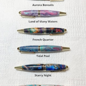 May include: Six pens with different colorful marbled patterns. The pens are all the same size and have gold accents. The pens are labeled with names like "Aurora Borealis", "Land of Many Waters", "French Quarter", "Tidal Pool", "Starry Night", and "Ocean Etude".