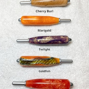 May include: Five crochet hook handles with silver metal tips, arranged vertically. The handles are labeled: Cherry Burl, Marigold, Twilight, Goldfish, and Apricot. Text at the top states: All handles are the same size. Each handle features a unique color and pattern, including wood grain, solid orange, purple and gold swirl, textured green and orange, and marbled red.