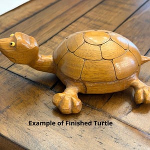 May include: A carved wooden turtle figurine with a smooth, light brown finish. The turtle features a segmented shell pattern, a small head with yellow eyes, and four distinct feet. The text "Example of Finished Turtle" is visible below.