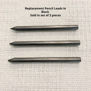 May include: Three black replacement pencil leads arranged horizontally. The text above the leads reads: Replacement Pencil Leads in Black. Sold in set of 3 pieces. Each lead is cylindrical with a pointed tip and a flat end.