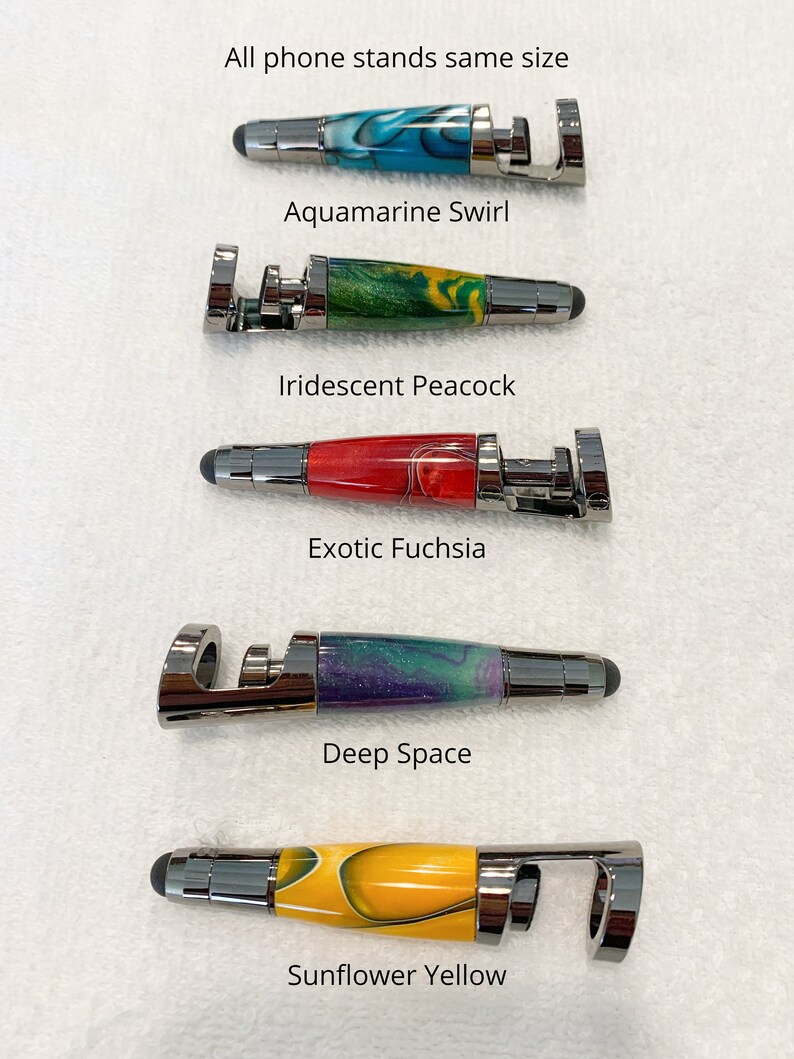 May include: Six phone stands with different colored wooden inlays. The stands are all the same size and have a metal ring on the top. The colors of the inlays are aquamarine swirl, iridescent peacock, exotic fuchsia, deep space, and sunflower yellow.