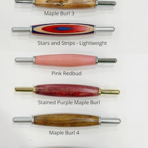 May include: Six seam rippers with wooden handles arranged vertically. The handles are labeled: Maple Burl 3, Stars and Strips, Pink Redbud, Stained Purple Maple Burl, Maple Burl 4, and Maple Burl 5. The metal tips are silver or gold colored.