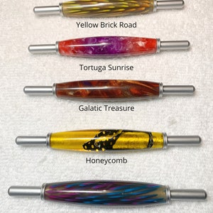 May include: Five 2-in-1 tools with cylindrical barrels and silver metal ends. The barrels feature distinct patterns: Yellow Brick Road, Tortuga Sunrise, Galatic Treasure, Honeycomb, and Night Music. Text labels identify each tool by name.