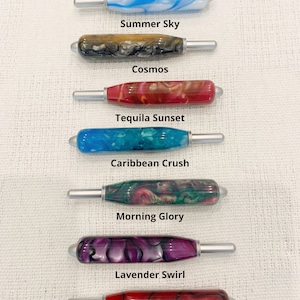 May include: Seven crochet hooks with marbled resin handles in various colors, including blue, black, red, teal, green, purple, and fire engine red. Each handle features a silver metal hook tip. Text labels identify the colors as Summer Sky, Cosmos, Tequila Sunset, Caribbean Crush, Morning Glory, Lavender Swirl, and Fire Engine Red Swirl. The text states all crochet handles are the same size.