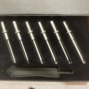 Op de afbeelding: Een set van zes zilverkleurige metalen gereedschappen met een zwarte handgreep, netjes gerangschikt in een zwarte doos met een fluwelen voering. De gereedschappen hebben een cilindrisch ontwerp met een puntig uiteinde. De doos is rechthoekig.