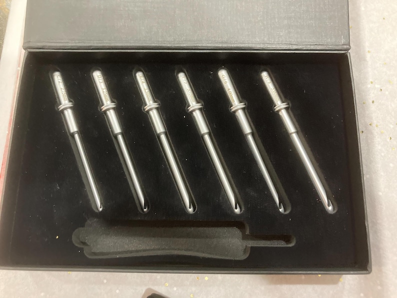 Op de afbeelding: Een set van zes zilverkleurige metalen gereedschappen met een zwarte handgreep, netjes gerangschikt in een zwarte doos met een fluwelen voering. De gereedschappen hebben een cilindrisch ontwerp met een puntig uiteinde. De doos is rechthoekig.