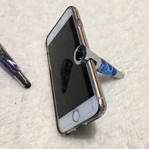 May include: Two stylus pens with a swirl design, one purple and one blue, with a silver metal top. The blue pen is propped up against a clear phone case on a white fuzzy surface.