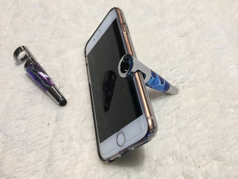 May include: Two stylus pens with a swirl design, one purple and one blue, with a silver metal top. The blue pen is propped up against a clear phone case on a white fuzzy surface.
