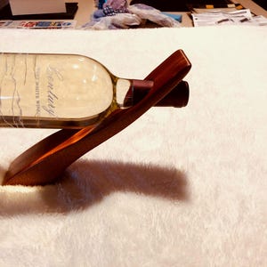 May include: A wooden wine bottle holder with a bottle of Century West White Wine. The holder is made of dark brown wood and has a curved design to cradle the bottle. The wine bottle is clear with a label.