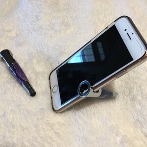 May include: A purple and silver pen with a ring on the top is holding up a white smartphone with a black screen. The phone is angled and the pen is standing upright.