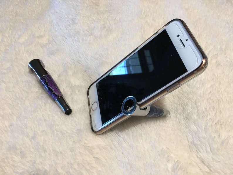 May include: A purple and silver pen with a ring on the top is holding up a white smartphone with a black screen. The phone is angled and the pen is standing upright.