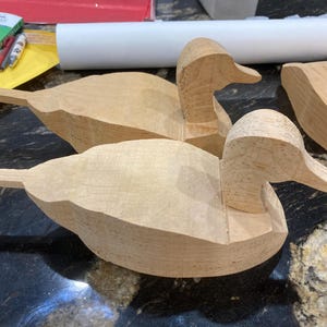 May include: Two unfinished wooden duck carvings. The ducks are light brown and have been roughly shaped, with details like the head and tail feathers visible. The ducks are on a dark surface.