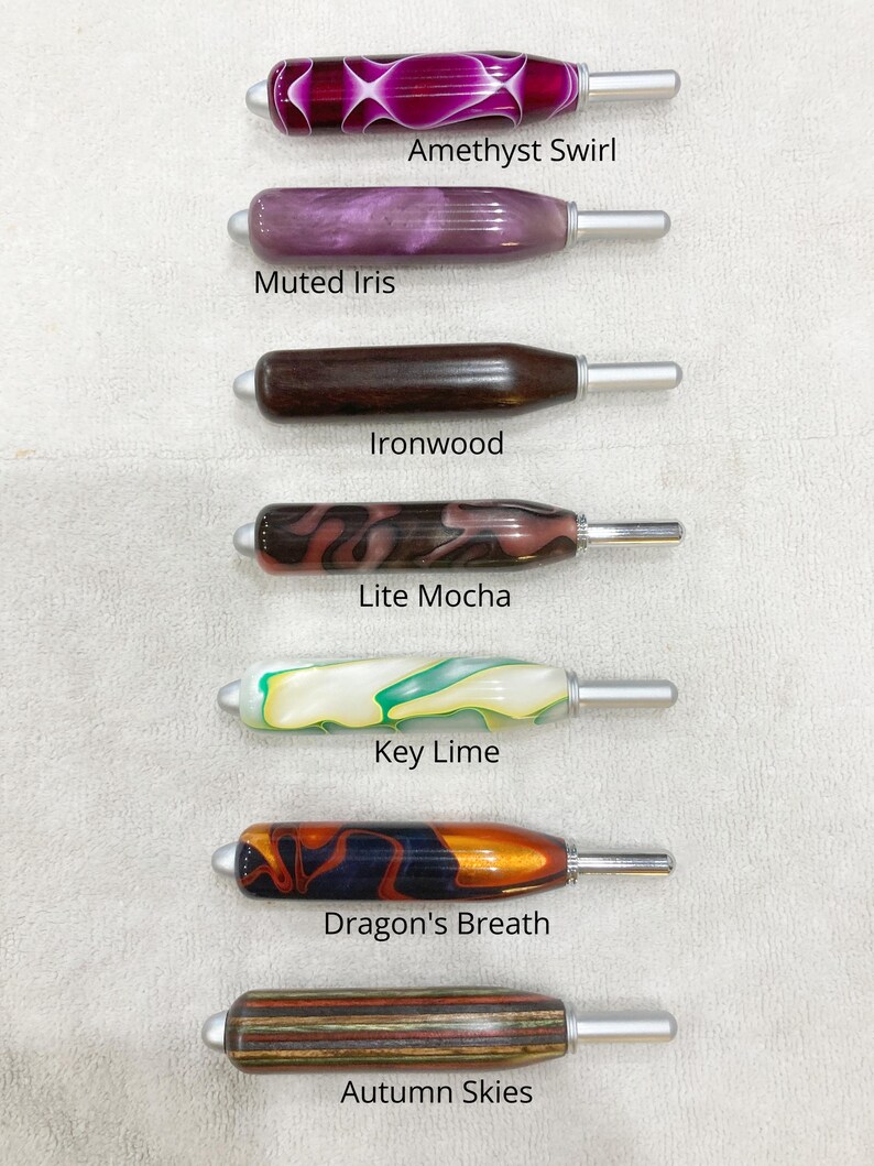 May include: Seven crochet hooks with ergonomic handles in various colors and patterns, labeled Amethyst Swirl, Muted Iris, Ironwood, Lite Mocha, Key Lime, Dragon's Breath, and Autumn Skies. Each hook features a smooth, rounded handle and a silver tip.