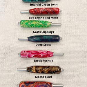 May include: Seven crochet hook handles with silver metal tips, each featuring a unique resin pattern: Emerald Green Swirl, Fire Engine Red Mesh, Grass Clippings, Deep Space, Exotic Fuchsia, Mocha Swirl, and Fiesta. All crochet handles are the same size.