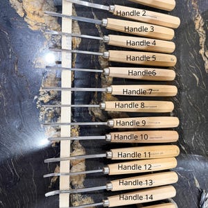 May include: A set of 15 wood carving chisels with wooden handles and metal blades. Each handle is labeled with a number from 1 to 15. The chisels are arranged vertically against a light-colored wooden support.