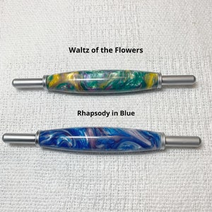 May include: Two crochet hooks with colorful marbled handles. The top hook has a green, yellow, and purple marbled handle and is labeled "Waltz of the Flowers". The bottom hook has a blue, white, and purple marbled handle and is labeled "Rhapsody in Blue".