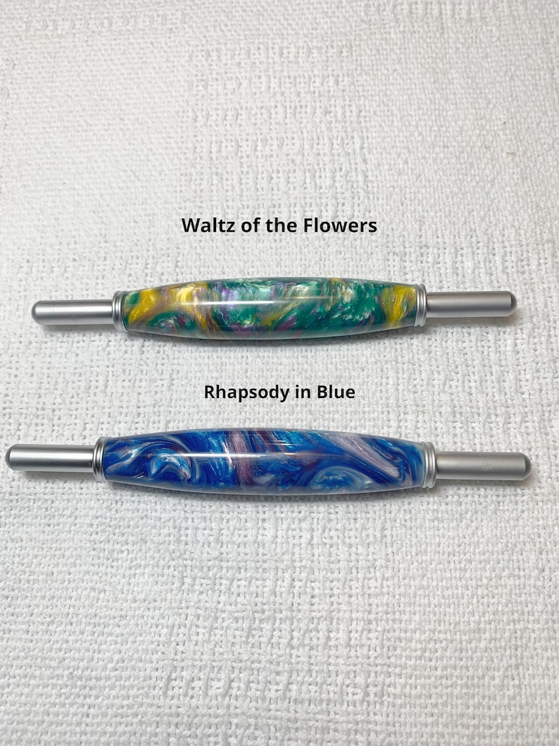 May include: Two crochet hooks with colorful marbled handles. The top hook has a green, yellow, and purple marbled handle and is labeled "Waltz of the Flowers". The bottom hook has a blue, white, and purple marbled handle and is labeled "Rhapsody in Blue".
