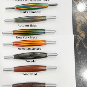 May include: Eight seam rippers with turned wooden handles in various colors and patterns, labeled Purple Stained Seam Ripper, God's Rainbow, Autumn Skies, New York Metz, Hawaiian Sunset, Tuxedo, Bloodwood, and Pheasantwood, each with metal tips.