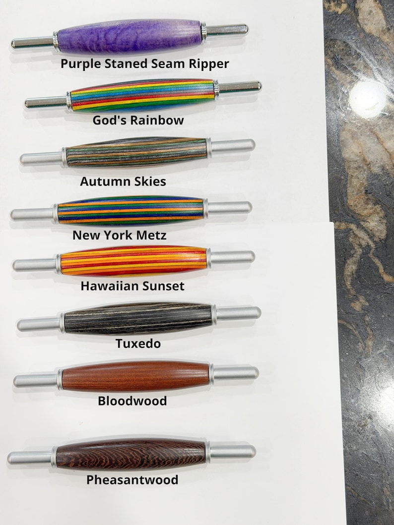 May include: Eight seam rippers with turned wooden handles in various colors and patterns, labeled Purple Stained Seam Ripper, God's Rainbow, Autumn Skies, New York Metz, Hawaiian Sunset, Tuxedo, Bloodwood, and Pheasantwood, each with metal tips.