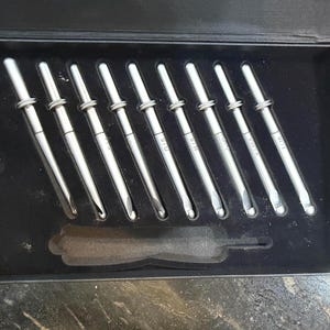 May include: A set of silver crochet hooks in a black box. The hooks are arranged neatly, with sizes labeled on each handle. A black foam insert holds the hooks securely. The box is open, revealing the contents.