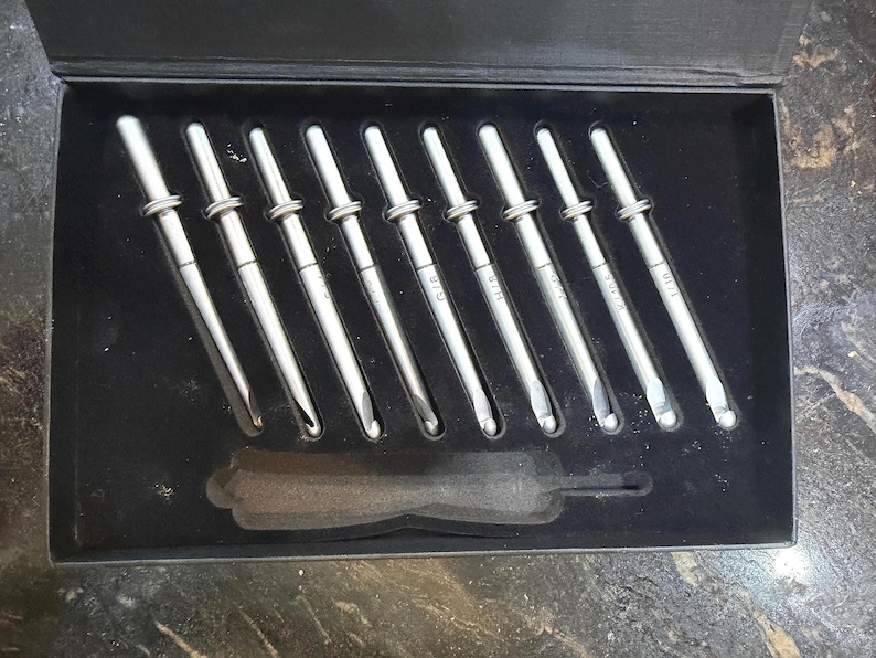 Op de afbeelding: Een set zilveren haaknaalden in een zwarte doos. De haaknaalden zijn netjes gerangschikt, met maten op elke handgreep. Een zwart schuimrubberen inzetstuk houdt de naalden op hun plaats. De doos is open en toont de inhoud.