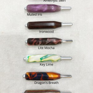 May include: Seven crochet hooks with ergonomic handles in various colors and patterns, labeled Amethyst Swirl, Muted Iris, Ironwood, Lite Mocha, Key Lime, Dragon's Breath, and Autumn Skies. Each hook features a smooth, rounded handle and a silver tip.