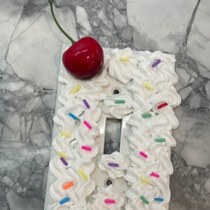 May include: White light switch cover decorated with white frosting and colorful sprinkles, topped with a red cherry.