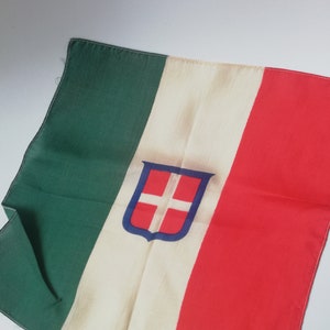 Può includere: Una bandiera rettangolare con strisce verticali verdi, bianche e rosse. Uno scudo blu e rosso con una croce bianca è centrato sulla striscia bianca. La bandiera è fatta di tessuto e ha un aspetto leggermente stropicciato.