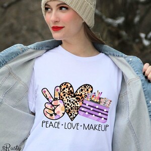 May include: White t-shirt with a peace sign, heart, and makeup bag design. The peace sign is made of makeup products, the heart is leopard print, and the makeup bag is purple with white polka dots. The text "Peace Love Makeup" is printed below the design.