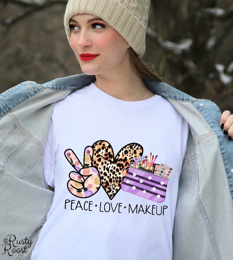 May include: White t-shirt with a peace sign, heart, and makeup bag design. The peace sign is made of makeup products, the heart is leopard print, and the makeup bag is purple with white polka dots. The text "Peace Love Makeup" is printed below the design.