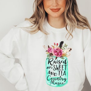 May include: White sweatshirt with a teal mason jar design featuring pink flowers and feathers. The jar has the text "Raised on Sweet and Tea Country Music".