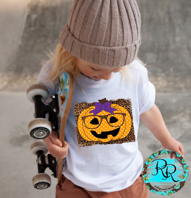 Puede incluir: Camiseta blanca con un gr&aacute;fico de una calabaza con gafas y un lazo morado sobre un fondo de estampado de leopardo. La calabaza tiene lunares blancos.
