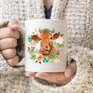 May include: White ceramic mug featuring a brown cow with a floral wreath design. The cow has its tongue out, surrounded by pink roses, succulents, and greenery. The mug is held by a person wearing a gray sweater.