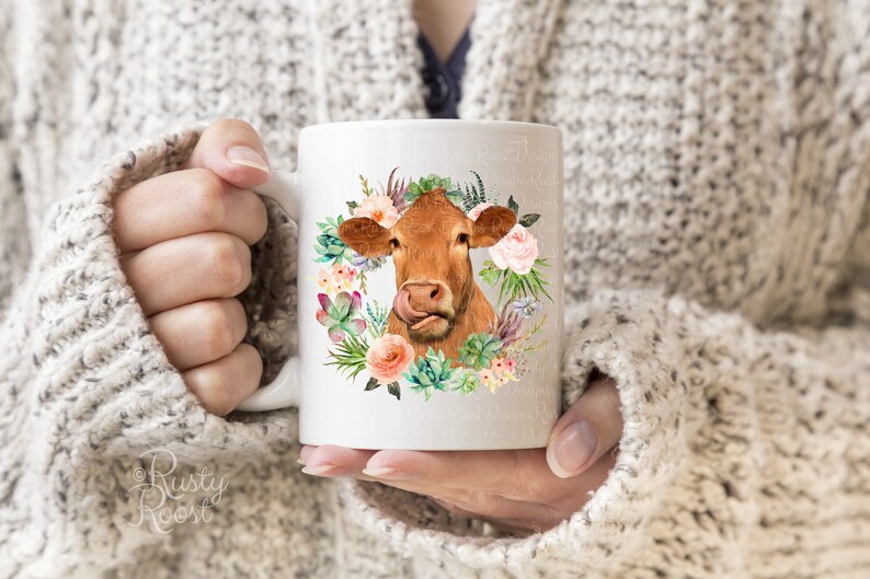 May include: White ceramic mug featuring a brown cow with a floral wreath design. The cow has its tongue out, surrounded by pink roses, succulents, and greenery. The mug is held by a person wearing a gray sweater.