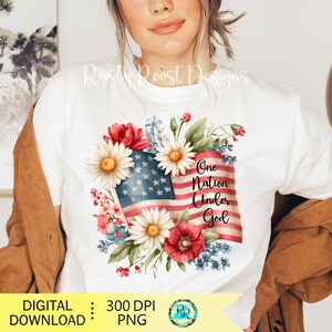 May include: White t-shirt featuring a watercolor design of the American flag, daisies, and floral accents. The text "One Nation Under God" is displayed on the flag. The design is surrounded by red, white, and blue flowers.