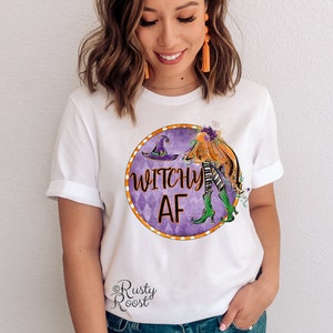 May include: White t-shirt with a purple and orange circle graphic featuring a witch with a pumpkin and the text "Witchy AF". The graphic is surrounded by a white and orange polka dot border. The text "Rusty Roost" is in the bottom left corner.