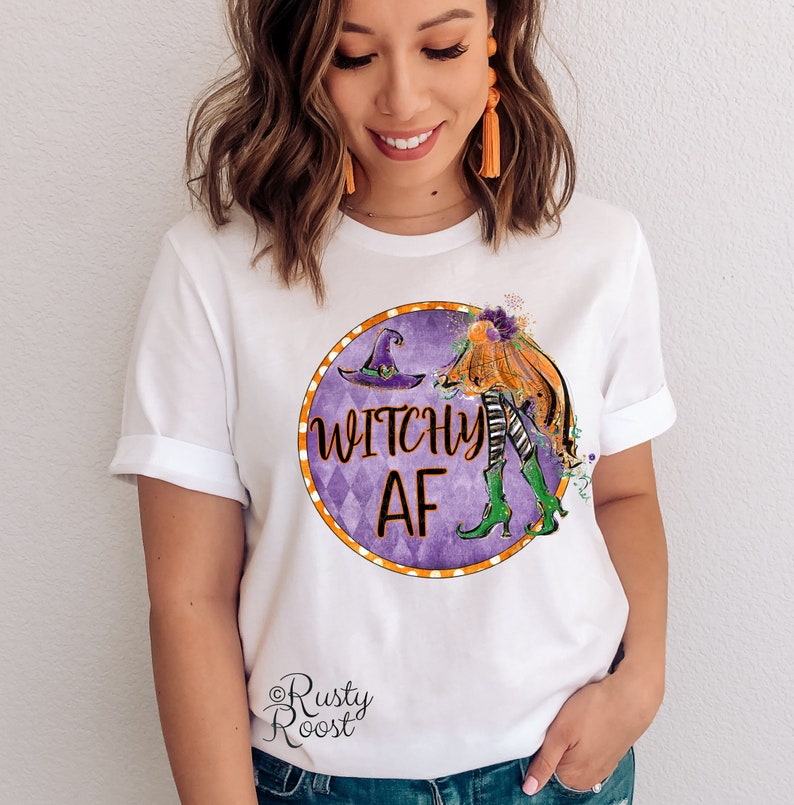 May include: White t-shirt with a purple and orange circle graphic featuring a witch with a pumpkin and the text "Witchy AF". The graphic is surrounded by a white and orange polka dot border. The text "Rusty Roost" is in the bottom left corner.