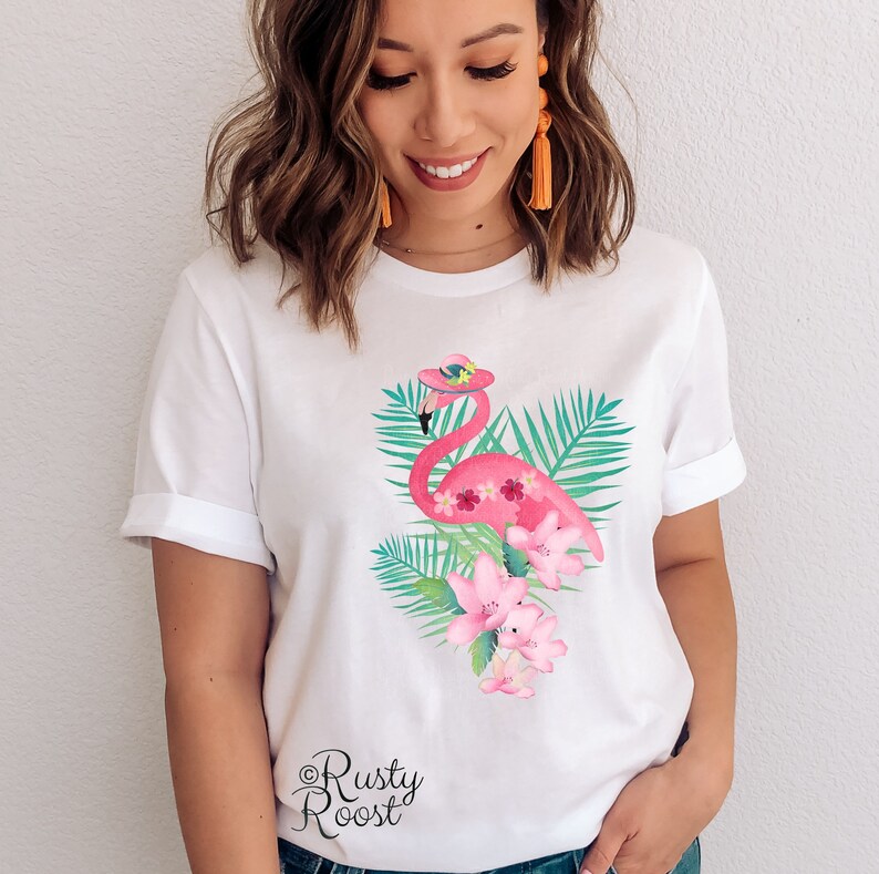 Peut inclure: T-shirt blanc avec un flamant rose portant un chapeau et entour&eacute; de feuilles de palmier vertes et de fleurs roses. Le texte "Rusty Roost" est imprim&eacute; en bas du t-shirt.