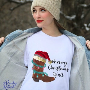 K&ouml;nnte beinhalten: Wei&szlig;es T-Shirt mit einem braunen Cowboy-Stiefel, der einen Weihnachtsmann-Hut und Weihnachtslichter tr&auml;gt. Der Text "Merry Christmas y'all" ist auf das T-Shirt gedruckt.