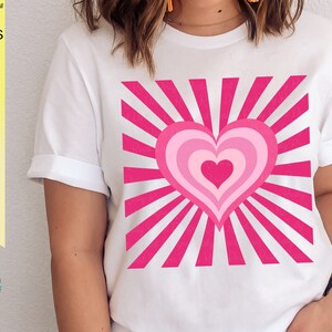 May include: A white t-shirt with a pink heart design. The heart is layered with concentric circles of pink and has a sunburst pattern behind it.