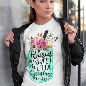 May include: White t-shirt with a teal mason jar graphic featuring flowers and feathers. The text on the jar reads "Raised on Sweet and Tea Country Music".
