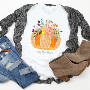 May include: A white t-shirt with a graphic of a pumpkin with a floral design. The pumpkin is orange and yellow with a honeycomb pattern. The flowers are pink, yellow, and green. The text "Rusty Roost Designs" is printed below the pumpkin.