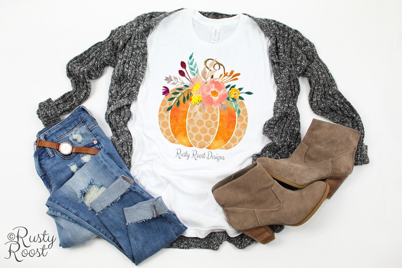 May include: A white t-shirt with a graphic of a pumpkin with a floral design. The pumpkin is orange and yellow with a honeycomb pattern. The flowers are pink, yellow, and green. The text "Rusty Roost Designs" is printed below the pumpkin.