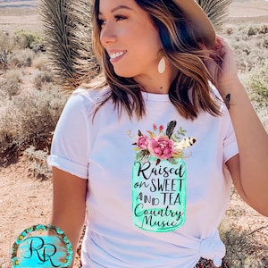 May include: White t-shirt with a teal mason jar graphic that says "Raised on Sweet and Tea Country Music". The jar is decorated with pink flowers and feathers.