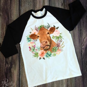 May include: A white raglan shirt with black sleeves. The shirt features a watercolor illustration of a brown cow's face, with its tongue sticking out, surrounded by a floral wreath of succulents and pink flowers.