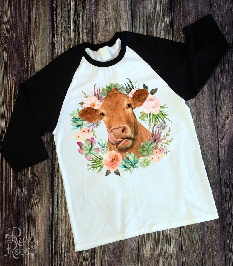 May include: A white raglan shirt with black sleeves. The shirt features a watercolor illustration of a brown cow's face, with its tongue sticking out, surrounded by a floral wreath of succulents and pink flowers.