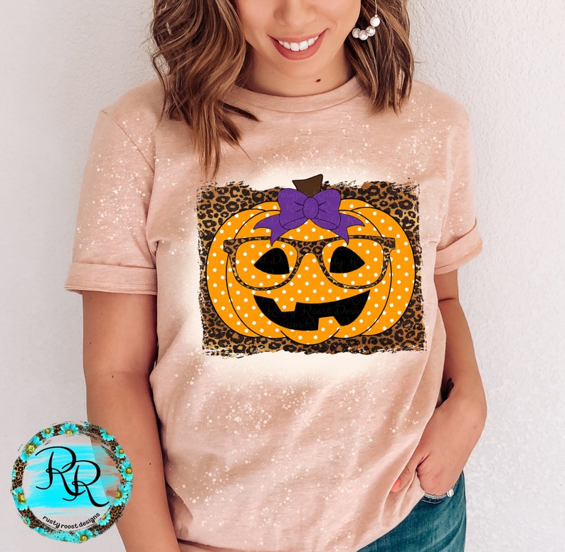 Puede incluir: Una camiseta rosa claro con un aspecto desgastado que presenta una calabaza con un lazo morado y gafas. La calabaza tiene lunares blancos y est&aacute; sobre un fondo de estampado de leopardo.