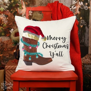 K&ouml;nnte beinhalten: Ein wei&szlig;es Kissen mit einem braunen Cowboy-Stiefel, der einen Weihnachtsmann-Hut tr&auml;gt und mit Weihnachtslichtern verziert ist. Der Text "Merry Christmas y'all" ist auf dem Kissen aufgedruckt.
