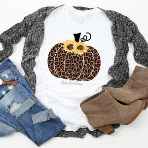 May include: White t-shirt with a brown and black leopard print pumpkin with yellow sunflowers for eyes. The text "Rusty Roost Designs" is printed below the pumpkin.