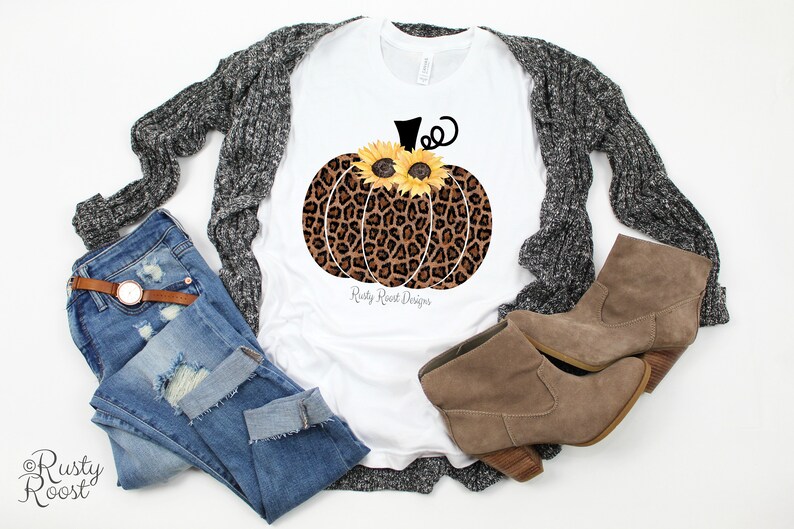 May include: White t-shirt with a brown and black leopard print pumpkin with yellow sunflowers for eyes. The text "Rusty Roost Designs" is printed below the pumpkin.